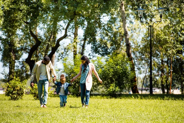 Parents afro-américains et fille se tenant la main et marchant sur l'herbe verte dans le parc — Photo de stock