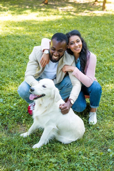 Sorridente casal afro-americano abraçando e agachamento perto do cão no parque — Fotografia de Stock