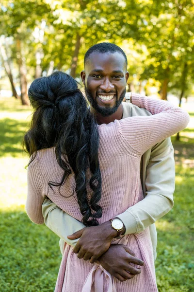 Sonriente afroamericano novio abrazando novia y mirando a la cámara en parque - foto de stock