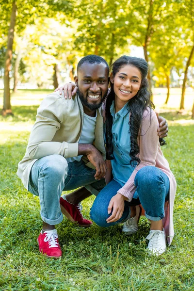 Africano americano pareja abrazos, sentadilla y mirando a cámara en parque - foto de stock