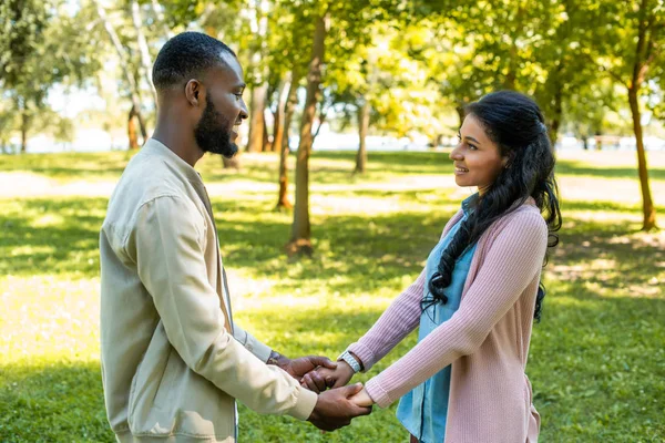 Sonriente africano americano pareja cogido de la mano y mirando el uno al otro en parque - foto de stock