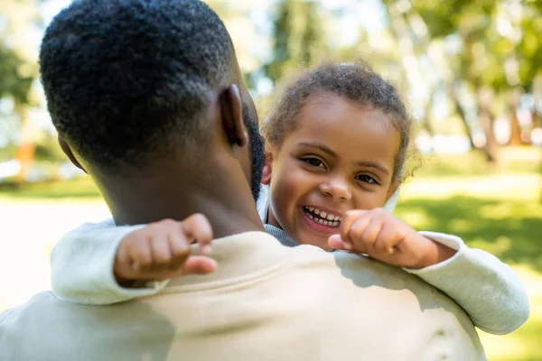 Счастливая африканская американская дочь смотрит в камеру, пока отец обнимает ее в парке — стоковое фото
