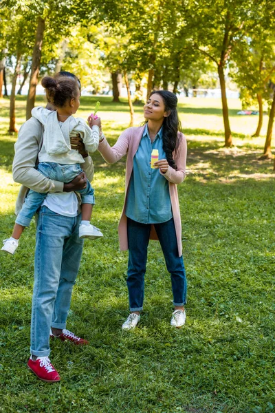 Afro-americana mãe ajudando filha soprando bolhas de sabão no parque — Fotografia de Stock