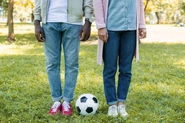 Imagen recortada de pareja afroamericana de pie cerca de la pelota de fútbol en el parque - foto de stock