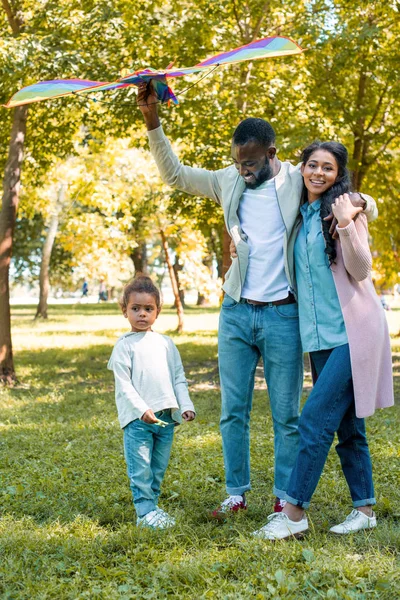 Afro-americano pai abraçando esposa e segurando papagaio acima filha no parque — Fotografia de Stock