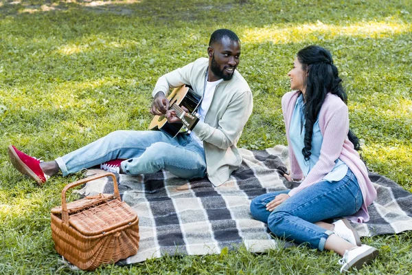 African american boyfriend playing acoustic guitar for girlfriend at picnic in park — Stock Photo