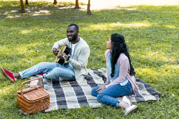 Smiling african american boyfriend playing acoustic guitar for girlfriend in park — Stock Photo