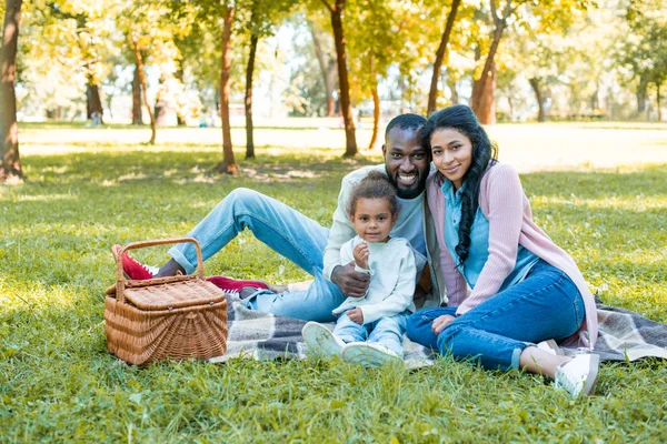 Sonrientes afroamericanos padres e hija sentados en el picnic y mirando a la cámara en el parque - foto de stock