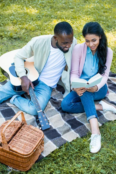 Africano americano namorado segurando guitarra acústica e namorada leitura livro no parque — Fotografia de Stock