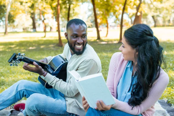 Sonriente afroamericano novio jugando guitarra acústica y novia lectura libro en parque - foto de stock