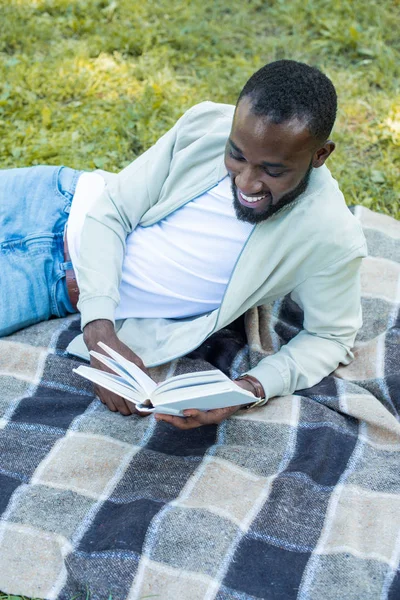 Happy handsome african american man lying on blanket in park and reading book — Stock Photo