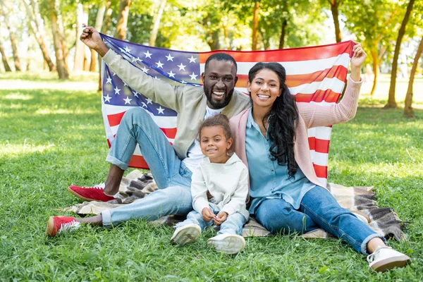 Sonrientes padres afroamericanos y su hija sosteniendo bandera americana en el picnic en el parque - foto de stock