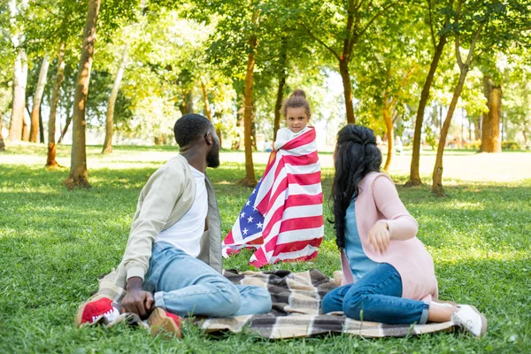 Hija afroamericana de pie envuelta en bandera americana en parque - foto de stock
