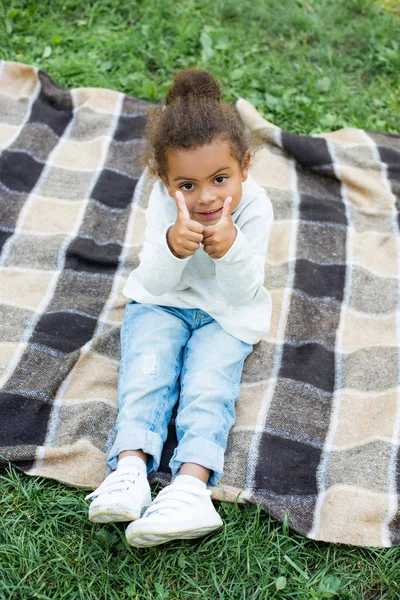 Adorable african american kid sitting on blanket and showing thumbs up in park — Stock Photo
