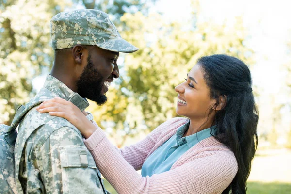 Vue de côté de sourire afro-américain soldat en uniforme militaire étreignant petite amie dans le parc — Photo de stock