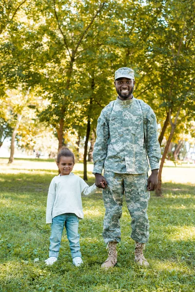 Sonriente soldado afroamericano en uniforme militar e hija tomados de la mano en el parque - foto de stock