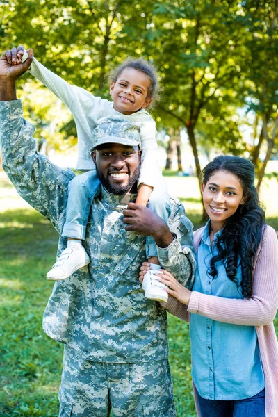 Sonriente afroamericano soldado en uniforme militar sosteniendo hija sobre hombros en parque - foto de stock