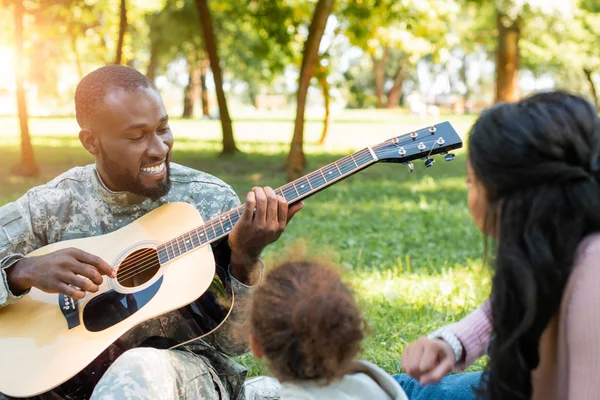 Felice soldato afroamericano in uniforme militare suonare la chitarra per la famiglia nel parco — Foto stock