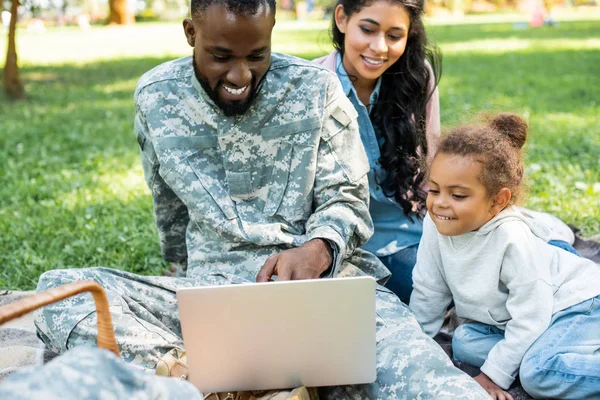 Sonriente soldado afroamericano en uniforme militar usando portátil con la familia en el parque - foto de stock