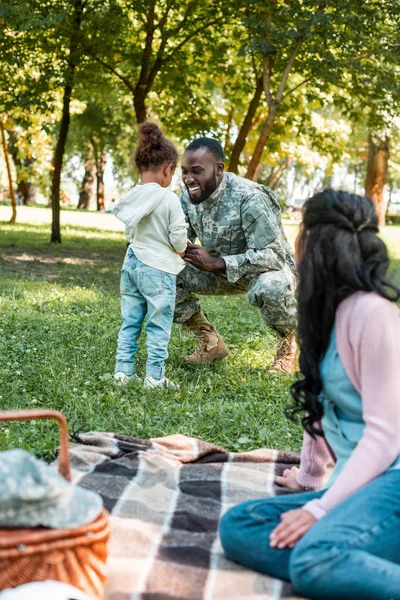 Улыбающийся африканский американский солдат присел рядом с дочерью в парке — стоковое фото