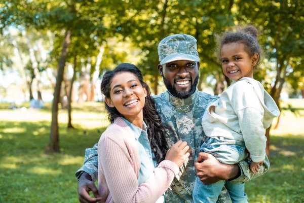 Felice soldato afroamericano in uniforme militare guardando la macchina fotografica con la famiglia nel parco — Foto stock