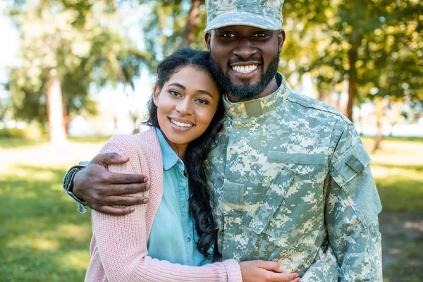 Sonriente afroamericano soldado en uniforme militar abrazando novia en parque - foto de stock