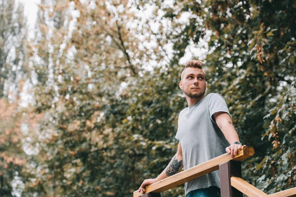 Jeune homme tatoué appuyé sur des balustrades dans le parc d'automne — Photo de stock