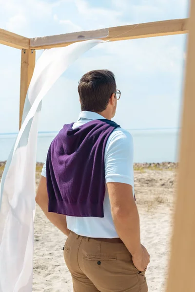 Vista trasera del hombre de pie cerca de la decoración con cortina blanca de encaje en la playa de arena - foto de stock