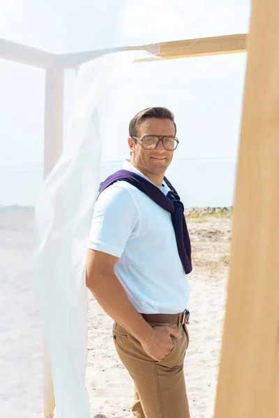 Hombre guapo en gafas de pie cerca de la decoración con cortina blanca de encaje en la playa de arena - foto de stock