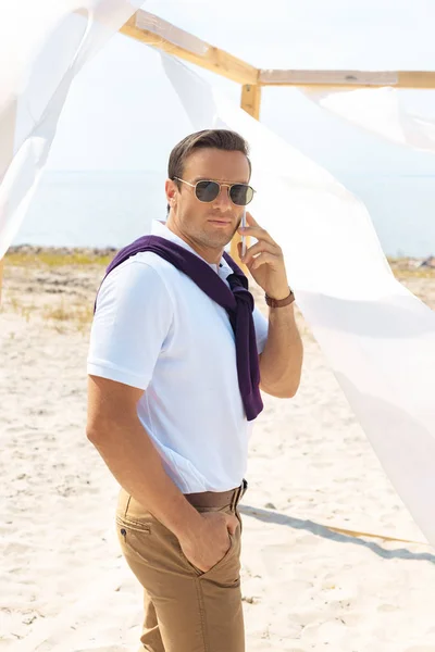 Vue latérale de l'homme élégant en lunettes de soleil parlant sur smartphone sur la plage de sable fin — Photo de stock