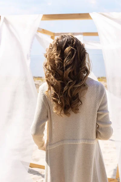 Back view of woman standing near wooden decoration with white curtain lace on sandy beach — Stock Photo