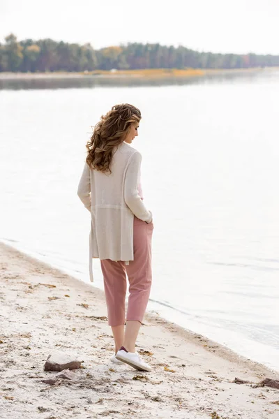 Vista lateral de la mujer pensativa con las manos en los bolsillos de pie en la playa de arena cerca del mar - foto de stock