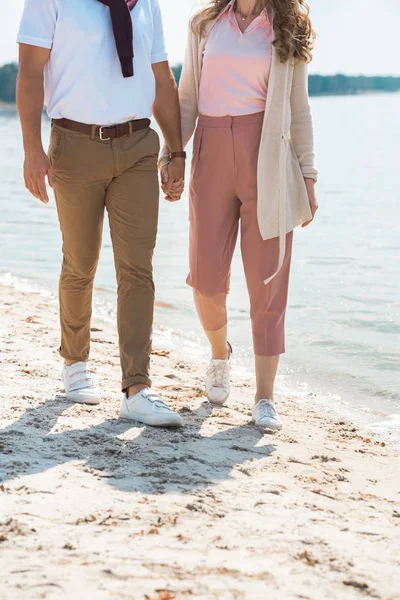 Vue partielle du couple romantique tenant la main tout en marchant sur le bord de la rivière sablonneuse — Photo de stock