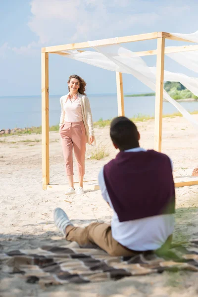 Selective focus of man on blanket looking at smiling wife walking on sandy beach — Stock Photo