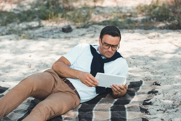 Hombre elegante que usa la tableta mientras descansa en la manta en el suelo arenoso - foto de stock