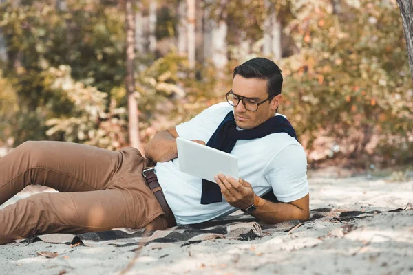 Hombre elegante que usa la tableta mientras descansa en la manta en el suelo arenoso - foto de stock