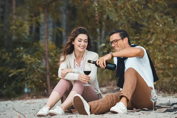 Homme verser du vin rouge dans le verre tout en se reposant avec femme sur la plage de sable — Photo de stock