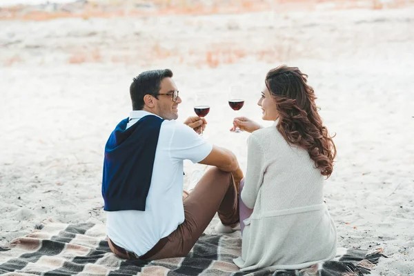 Visão traseira do casal com copos de vinho tinto descansando sobre cobertor na praia de areia — Fotografia de Stock