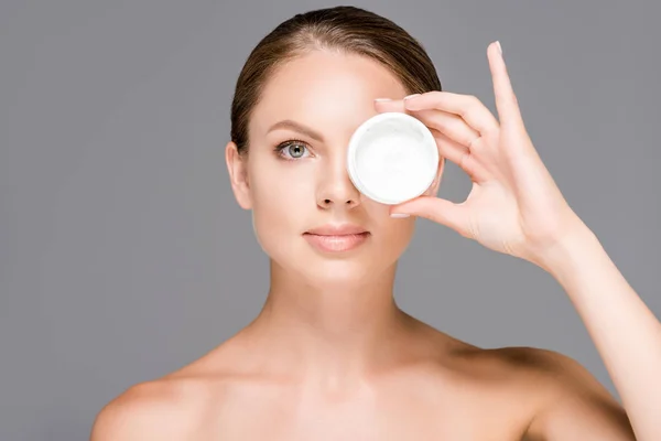 Portrait of beautiful young woman covering eye with face cream in jar isolated on grey — Stock Photo