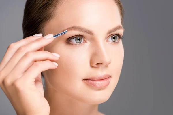 Portrait of young woman plucking eyebrows with tweezers isolated on grey — Stock Photo