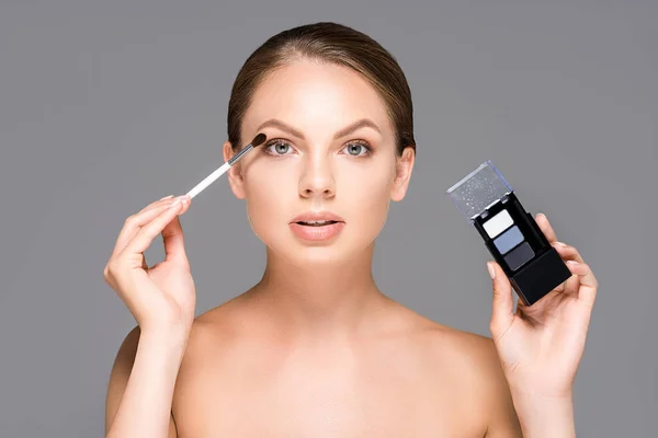 Retrato de mujer atractiva con sombras de ojos y cepillo de maquillaje aislado en gris - foto de stock