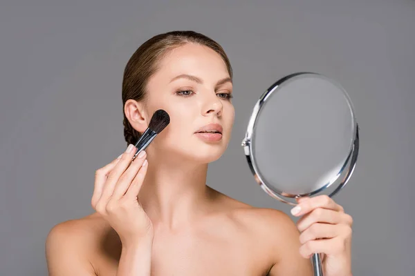 Retrato de jovem mulher olhando para o espelho e aplicando blush isolado no cinza — Fotografia de Stock