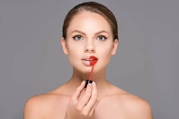 Retrato de una hermosa joven con hombros desnudos aplicando lápiz labial rojo aislado en gris - foto de stock