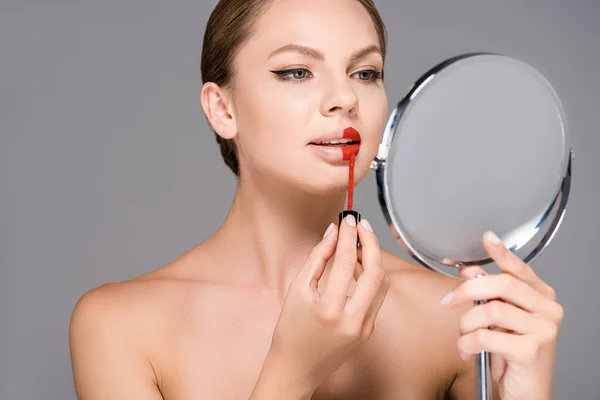 Ritratto di donna attraente che si guarda allo specchio e applica rossetto rosso isolato su grigio — Foto stock