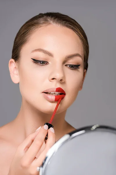 Porträt einer attraktiven Frau, die in den Spiegel schaut und roten Lippenstift isoliert auf grau aufträgt — Stockfoto