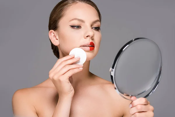 Portrait of young woman with red lipstick on half of lips and sponge looking at mirror isolated on grey — Stock Photo