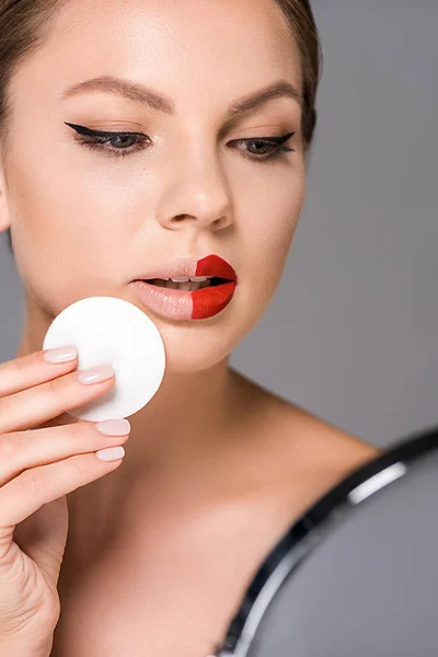 Retrato de jovem mulher com batom vermelho na metade dos lábios e esponja olhando espelho isolado no cinza — Fotografia de Stock