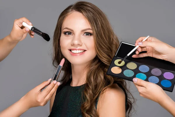 Partial view of makeup artists with cosmetics and beautiful smiling woman isolated on grey — Stock Photo