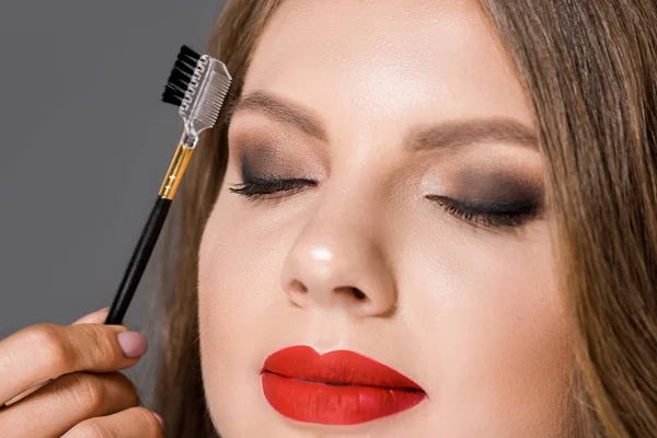 Partial view of makeup artist with brush for eyebrows and model with eyes closed isolated on grey — Stock Photo