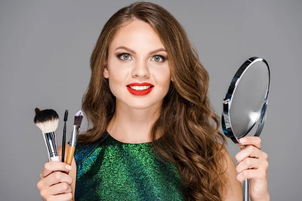 Portrait of beautiful smiling woman with makeup brushes and mirror isolated on grey — Stock Photo